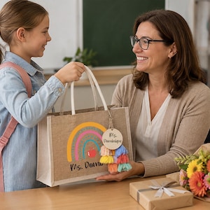 May include: A rectangular jute tote bag featuring a colorful rainbow graphic, a red apple icon, and the text "Ms. Dominique" printed on the front. A round wooden tag with the text "Ms. Dominique" and a multi-colored tassel are attached to the handle.