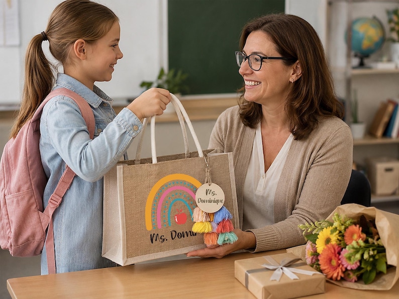 May include: A rectangular jute tote bag featuring a colorful rainbow graphic, a red apple icon, and the text "Ms. Dominique" printed on the front. A round wooden tag with the text "Ms. Dominique" and a multi-colored tassel are attached to the handle.