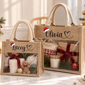 May include: Two rectangular burlap tote bags with clear plastic windows. The smaller bag features the name Lacey and a Santa hat, while the larger bag features the name Olivia and a Santa hat. Both bags contain wrapped gifts, greenery, and ornaments.