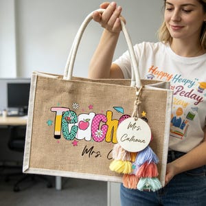 May include: A rectangular tan burlap tote bag with white fabric handles. The front features the word Teacher in colorful patterned letters with an apple and stars. A round wooden tag labeled Mrs. Calhoun hangs with a multi-colored tassel.