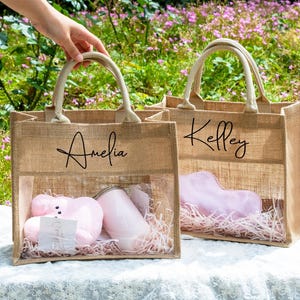 Könnte beinhalten: Zwei Jute-Tragetaschen mit transparenten Frontplatten, jede mit rosa Artikeln und dekorativem Stroh. Eine Tasche ist mit "Amelia" beschriftet, die andere mit "Kelley". Beide haben beige Griffe und stehen vor Grün und Blumen.