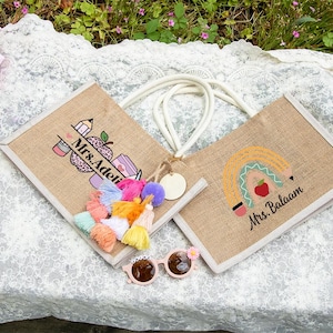 May include: Two rectangular jute tote bags with white rope handles. One bag features a pencil and apple graphic with the text Mrs. Adeli, while the other displays a rainbow and apple design with the text Mrs. Balaam. A colorful tassel charm is attached.
