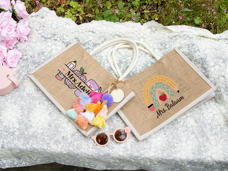 May include: Two rectangular jute tote bags with white rope handles. One bag features a pencil and apple graphic with the text Mrs. Adeli, while the other displays a rainbow and apple design with the text Mrs. Balaam. A colorful tassel charm is attached.