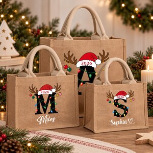 Op de afbeelding: Drie beige jute draagtassen met twee handvatten. Elke tas is voorzien van een grote zwarte letter versierd met kleurrijke kerstverlichting, een rendiergewei, een rode kerstmuts en het jaartal 2026. Twee tassen hebben namen eronder.