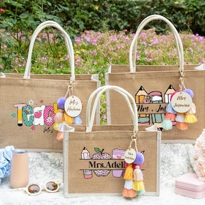 May include: Three rectangular jute tote bags with white handles, each featuring a personalized wooden tag and colorful tassel. The bags display printed graphics of pencils, apples, and text including Mrs. Helena, Mrs. Jasmine, and Mrs. Adeline.