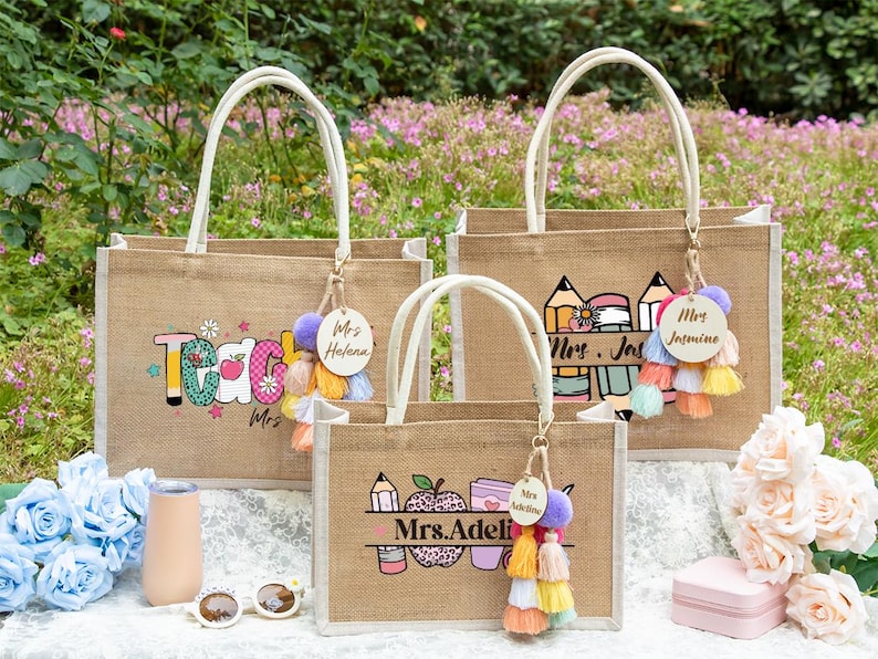 May include: Three rectangular jute tote bags with white handles, each featuring a personalized wooden tag and colorful tassel. The bags display printed graphics of pencils, apples, and text including Mrs. Helena, Mrs. Jasmine, and Mrs. Adeline.