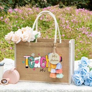 May include: A rectangular jute tote bag with white handles, featuring the word Teacher in colorful patterned letters and a small apple graphic. A round wooden keychain with the text Mrs. Helena hangs from a multi-colored tassel charm.