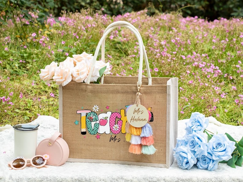May include: A rectangular jute tote bag with white handles, featuring the word Teacher in colorful patterned letters and a small apple graphic. A round wooden keychain with the text Mrs. Helena hangs from a multi-colored tassel charm.