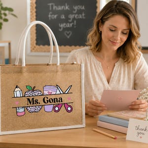 May include: A rectangular jute tote bag with white handles featuring a printed graphic of a pencil, a leopard print apple, a coffee cup, and scissors. The text "Ms. Gona" is printed in black across the center of the graphic design.