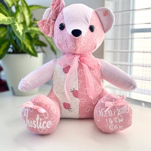May include: Pink and white patchwork teddy bear with a pink bow. The bear has a button nose and black eyes. The bear's paws are embroidered with the name "Justice" and the date "10/13/2020", weight "1 lb 7 oz", and length "12.6 in".