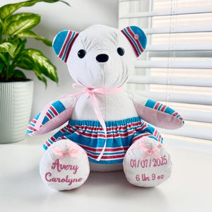 May include: White teddy bear with pink and blue striped fabric. The bear has a pink ribbon around its neck and pink bows on its feet. The bear's feet are embroidered with the text "Avery Caroline" and "01/07/2025 6 lbs 9 oz".