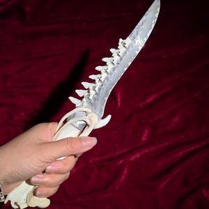 Puede incluir: Un cuchillo artesanal con mango de hueso y hoja dentada. La hoja tiene un patrón único y el mango está intrincadamente tallado. El cuchillo se muestra sobre un fondo rojo oscuro, resaltando su artesanía detallada.