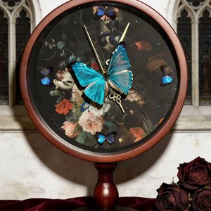 Puede incluir: Un reloj decorativo con una esfera floral oscura y un marco de madera. El reloj presenta una gran mariposa azul vibrante y varias mariposas más pequeñas. Un pájaro dorado corona el reloj. El reloj está montado sobre un soporte de madera.