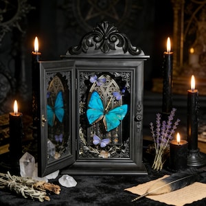 Puede incluir: Un expositor negro con una gran mariposa turquesa y varias mariposas moradas más pequeñas. El expositor está abierto, revelando un diseño de arco gótico. Velas encendidas y cristales rodean la exhibición, creando un ambiente místico.