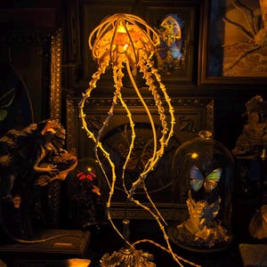 May include: A unique jellyfish-shaped lamp with a warm, golden glow. The lamp features intricate, branch-like tendrils and a decorative base. The background includes framed artwork and other decorative objects, creating a gothic or Victorian aesthetic.