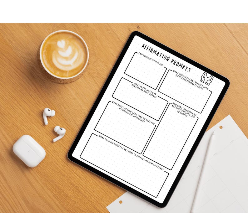May include: A digital tablet with the text "AFFIRMATION PROMPTS" and prompts for self-reflection. A cup of coffee with latte art, wireless earbuds, and a charging case are on a wooden surface.