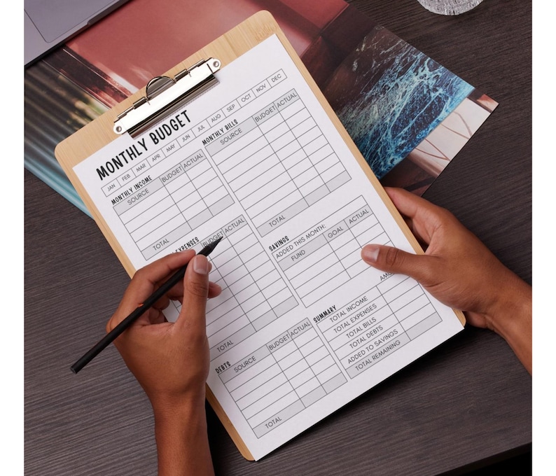 May include: A wooden clipboard with a metal clip holding a printed "MONTHLY BUDGET" form. The form has sections for income, bills, expenses, and savings. A black pen is held over the form.