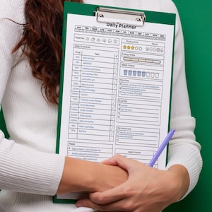 May include: A green clipboard with a "Daily Planner" sheet. The planner includes sections for daily schedules, priorities, and a habit tracker. A purple pen rests on the sheet. The person is wearing a white sweater and blue jeans.