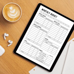 May include: A digital tablet displays a "MONTHLY BUDGET" template with sections for income, expenses, and bills. A cup of coffee with latte art, wireless earbuds, and a charging case are on a wooden surface.