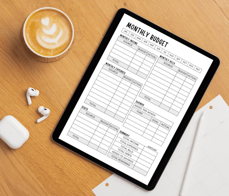 May include: A digital tablet displays a "MONTHLY BUDGET" template with sections for income, expenses, and bills. A cup of coffee with latte art, wireless earbuds, and a charging case are on a wooden surface.