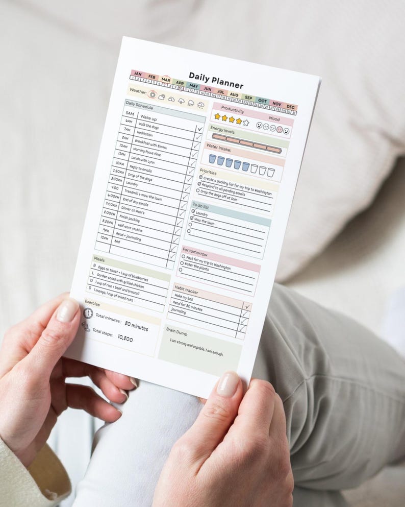 May include: A Daily Planner sheet with a colorful layout, held by a person. The planner includes sections for daily schedules, productivity, energy levels, mood, priorities, and a habit tracker. The text "Daily Planner" is at the top.