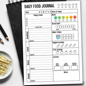 May include: A black spiral-bound notebook with a white page titled "DAILY FOOD JOURNAL." The journal includes sections for daily goals, mood, stress level, water intake, veggie and fruit servings, exercise, and notes. A pencil and gold paper clips are visible.