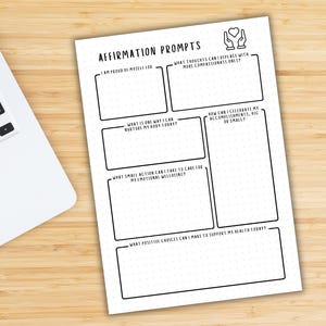 May include: A white affirmation prompts sheet with black text and a dotted grid pattern. The sheet includes prompts such as "I am proud of myself for" and "What thoughts can I replace with more compassionate ones?" A laptop is partially visible in the background.