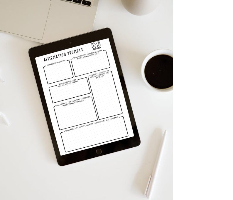 May include: A black tablet displaying an "Affirmation Prompts" template with prompts for self-reflection. The tablet is on a white surface, next to a laptop, a cup of coffee, and a pen. The template includes fill-in boxes for journaling.