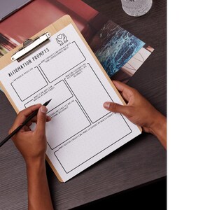 May include: A wooden clipboard with a silver clip holding a white paper with the text "Affirmation Prompts". The paper has boxes and prompts for writing. A black pen is held over the paper. The clipboard is on a dark surface.