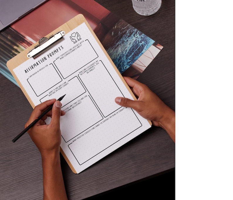 May include: A wooden clipboard with a silver clip holding a white paper with the text "Affirmation Prompts". The paper has boxes and prompts for writing. A black pen is held over the paper. The clipboard is on a dark surface.