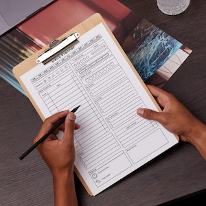 May include: A wooden clipboard with a silver clip holding a daily planner sheet. The planner includes sections for time, tasks, and mood tracking. A black pen is held over the planner, ready to write. The words "Daily Planner" are printed at the top.