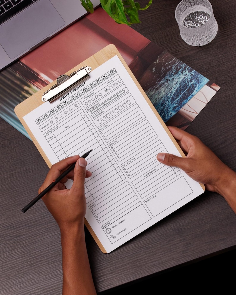May include: A wooden clipboard with a silver clip holding a daily planner sheet. The planner includes sections for time, tasks, and mood tracking. A black pen is held over the planner, ready to write. The words "Daily Planner" are printed at the top.