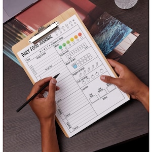 May include: A wooden clipboard with a printed "DAILY FOOD JOURNAL" form. The form includes sections for tracking mood, sleep, water intake, and food consumption. A black pen is held over the form, ready to write.