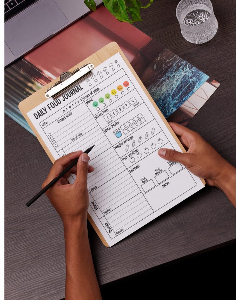 May include: A wooden clipboard with a printed "DAILY FOOD JOURNAL" form. The form includes sections for tracking mood, sleep, water intake, and food consumption. A black pen is held over the form, ready to write.