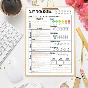 May include: A clipboard with a "Daily Food Journal" form, a cup of coffee, a computer mouse, and a keyboard. The form includes sections for goals, mood, water intake, and meal tracking. Gold paper clips, pencils, and a lipstick are also visible.