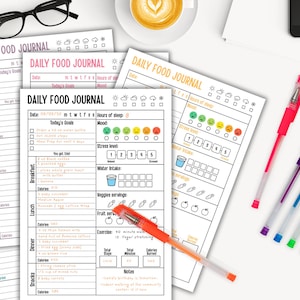 May include: A collection of printed daily food journals with colorful pens and a cup of coffee. The journals have sections for tracking meals, mood, water intake, and exercise. The text "DAILY FOOD JOURNAL" is visible on the pages.