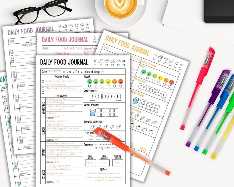 May include: A collection of printed daily food journals with colorful pens and a cup of coffee. The journals have sections for tracking meals, mood, water intake, and exercise. The text "DAILY FOOD JOURNAL" is visible on the pages.