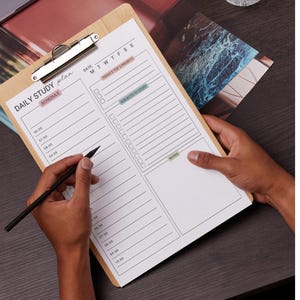 May include: A wooden clipboard with a metal clip holding a white paper with a "DAILY STUDY PLAN" layout. The plan includes sections for schedule, priorities, subjects to study, and notes. A black pen is held over the paper.