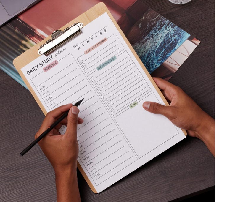 May include: A wooden clipboard with a metal clip holding a white paper with a "DAILY STUDY PLAN" layout. The plan includes sections for schedule, priorities, subjects to study, and notes. A black pen is held over the paper.