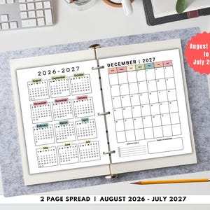 May include: A two-page spread calendar for August 2026 to July 2027, open on a gray felt surface. The left page displays a yearly overview, while the right page shows a monthly calendar for December 2027. A pencil rests below.