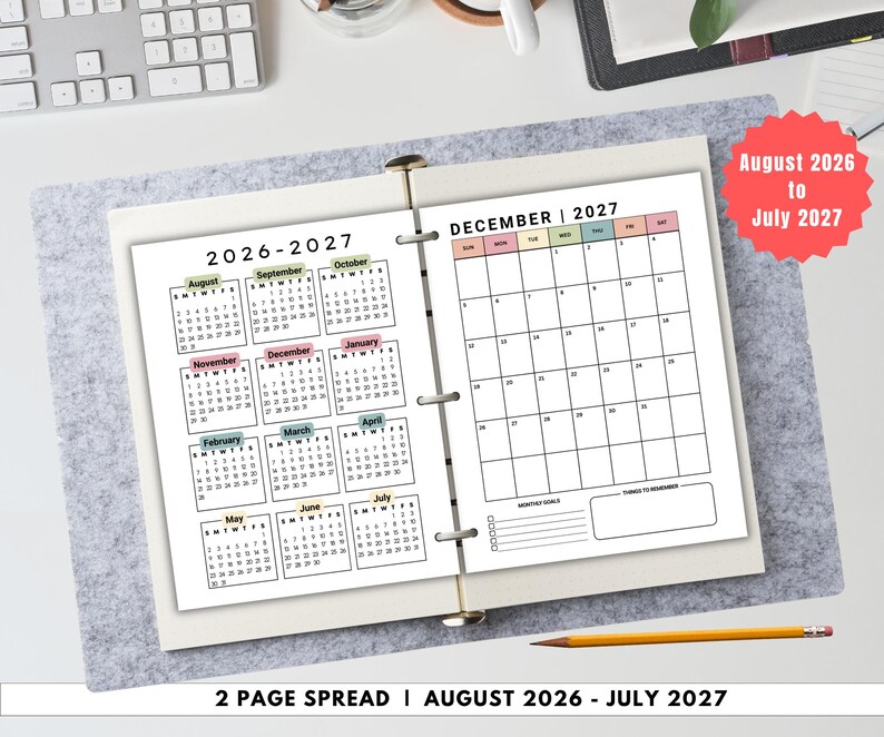 May include: A two-page spread calendar for August 2026 to July 2027, open on a gray felt surface. The left page displays a yearly overview, while the right page shows a monthly calendar for December 2027. A pencil rests below.