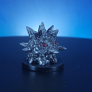 May include: A detailed, metallic-looking sculpture of a spiky, star-shaped object with two red gem-like eyes. The sculpture sits on a small, round, black base. The background is a gradient of blue.