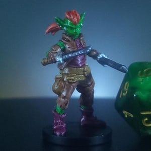 May include: A miniature fantasy figure of a green-skinned goblin with red hair, wearing brown and purple clothing, holding two swords. A large green die with white numbers is in the background. The figure is on a black base.