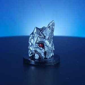 May include: A detailed, silver-colored fantasy figurine with a rough, textured surface. The figurine has a prominent, red, spherical eye and is mounted on a black, circular base. The background is a gradient of blue.