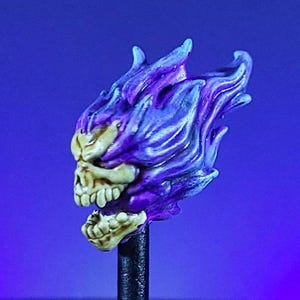 Puede incluir: Una calavera detallada, pintada a mano, con llamas moradas. La calavera es blanquecina con dientes visibles y una mandíbula detallada. Las llamas son un degradado de morado y azul, creando un efecto espectral. La calavera está montada sobre una varilla negra.