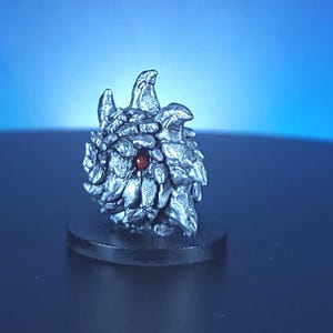 May include: A detailed, silver-colored sculpture of a creature's head, possibly a dragon or mythical beast, with textured scales and a prominent red gemstone eye. The sculpture is mounted on a small, round, black base.