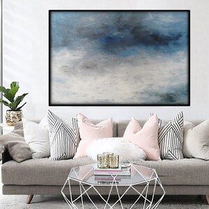 Op de afbeelding: Een groot abstract schilderij in blauw en wit. Het schilderij hangt aan een witte muur boven een grijze bank met kussens.