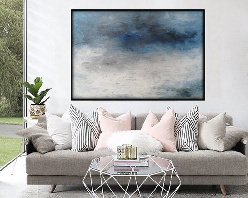 Op de afbeelding: Een groot abstract schilderij in blauw en wit. Het schilderij hangt aan een witte muur boven een grijze bank met kussens.