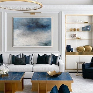 Op de afbeelding: Een witte bank met blauwe kussens staat in een woonkamer met een groot abstract schilderij van blauwe en witte wolken. Twee gouden en blauwe salontafels staan voor de bank. Een gouden plank met decoratieve items bevindt zich aan de rechterkant van de kamer.