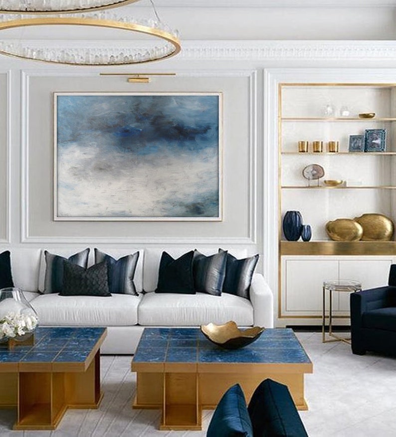 Op de afbeelding: Een witte bank met blauwe kussens staat in een woonkamer met een groot abstract schilderij van blauwe en witte wolken. Twee gouden en blauwe salontafels staan voor de bank. Een gouden plank met decoratieve items bevindt zich aan de rechterkant van de kamer.